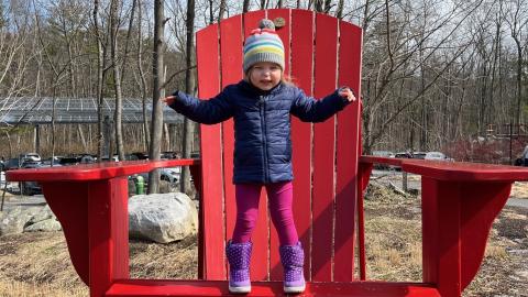 a small girl stand on the seat of a giant red Adirondack chair